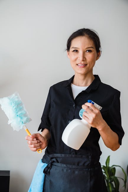A professional cleaner wearing red overalls and a gray t-shirt is operating a yellow upright vacuum cleaner on a light-colored tiled floor in a modern dining area. The cleaner is holding the vacuum's