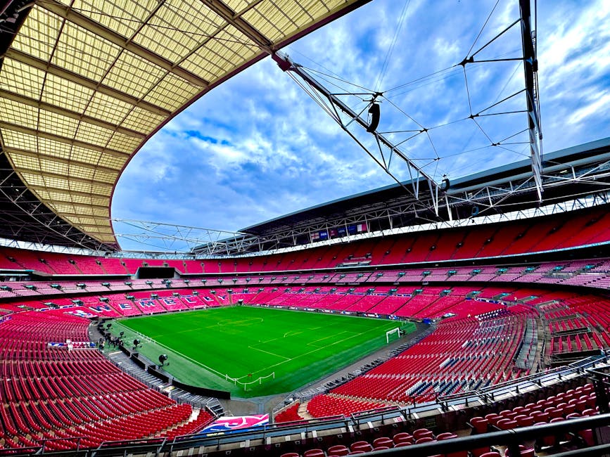 A panoramic view inside Wembley Stadium showing a grass football pitch surrounded by tiered seating filled with spectators. The pitch is green and well-maintained, with visible white markings and goal